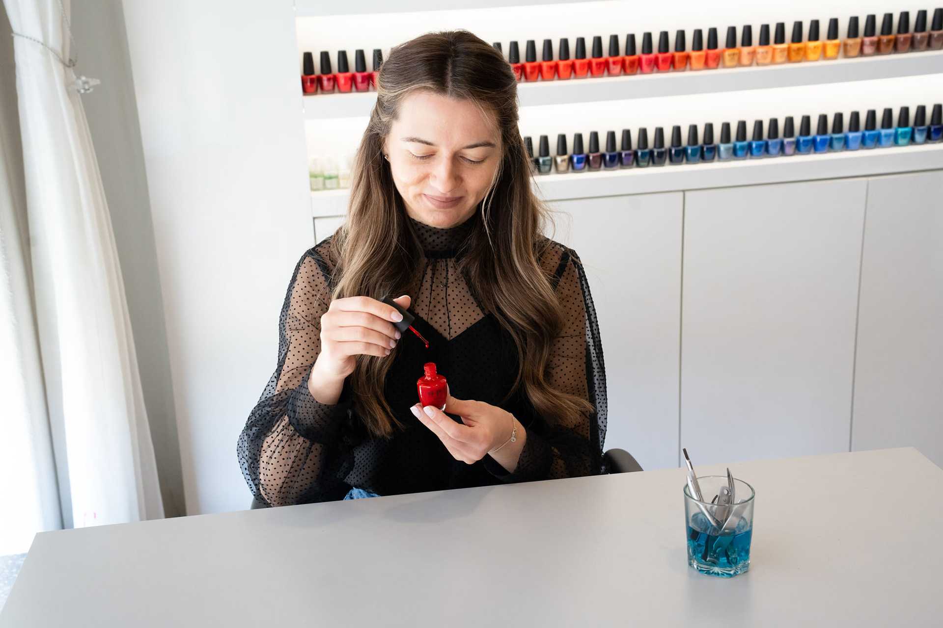 Woman painting nails with red polish at a salon, colorful bottles on shelf behind.
