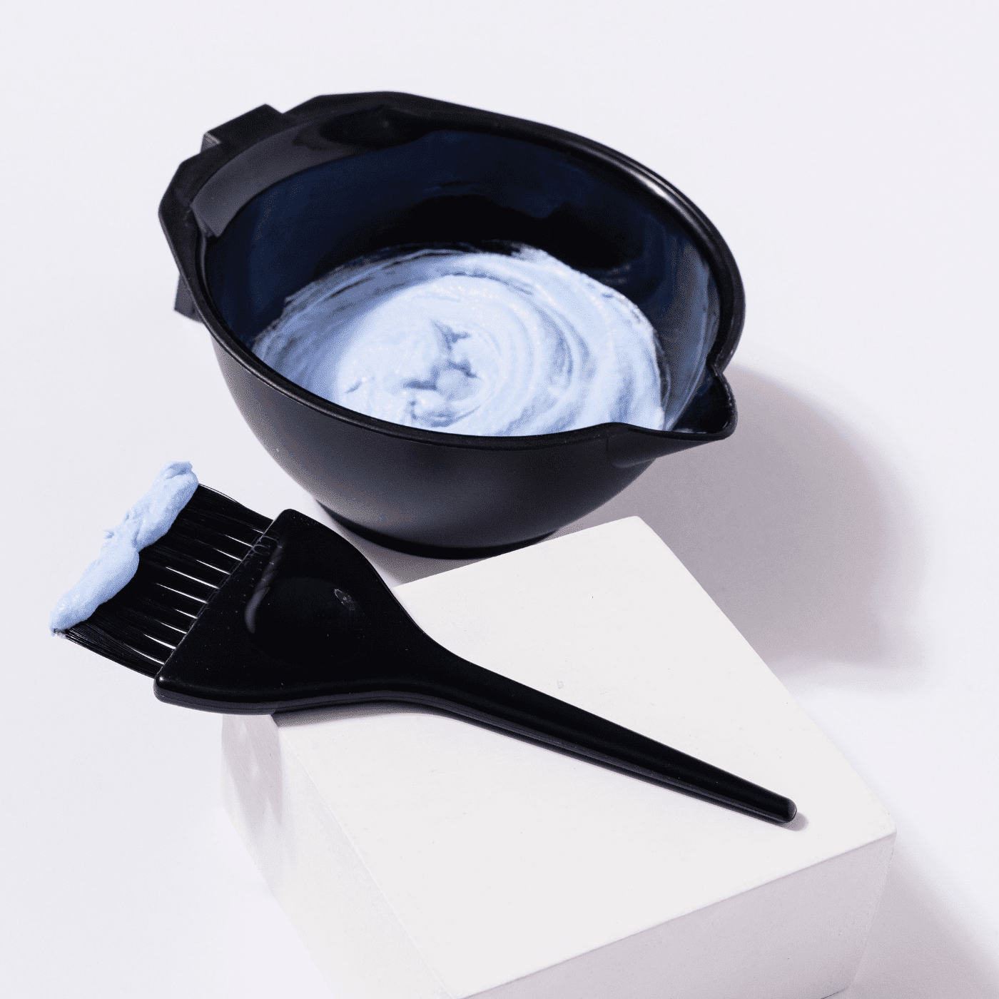 Black bowl with blue cream and brush on a white cube, used for hair dyeing.