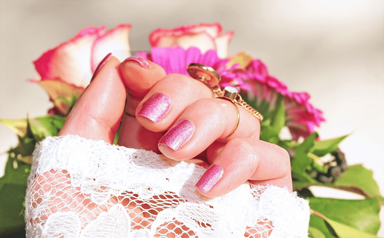 Hand with pink glitter nails holding flowers, adorned with lace and gold rings.